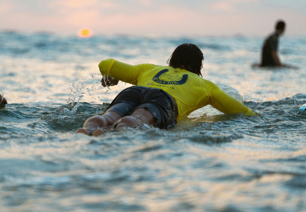 Surfer in yellow jersey paddles through ocean waves at sunset, Tel Aviv.